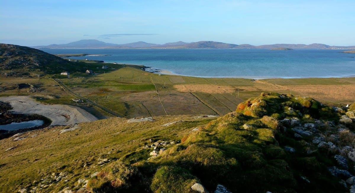 An elevated view over green fields and houses towards a white-sand bay and distant hills.