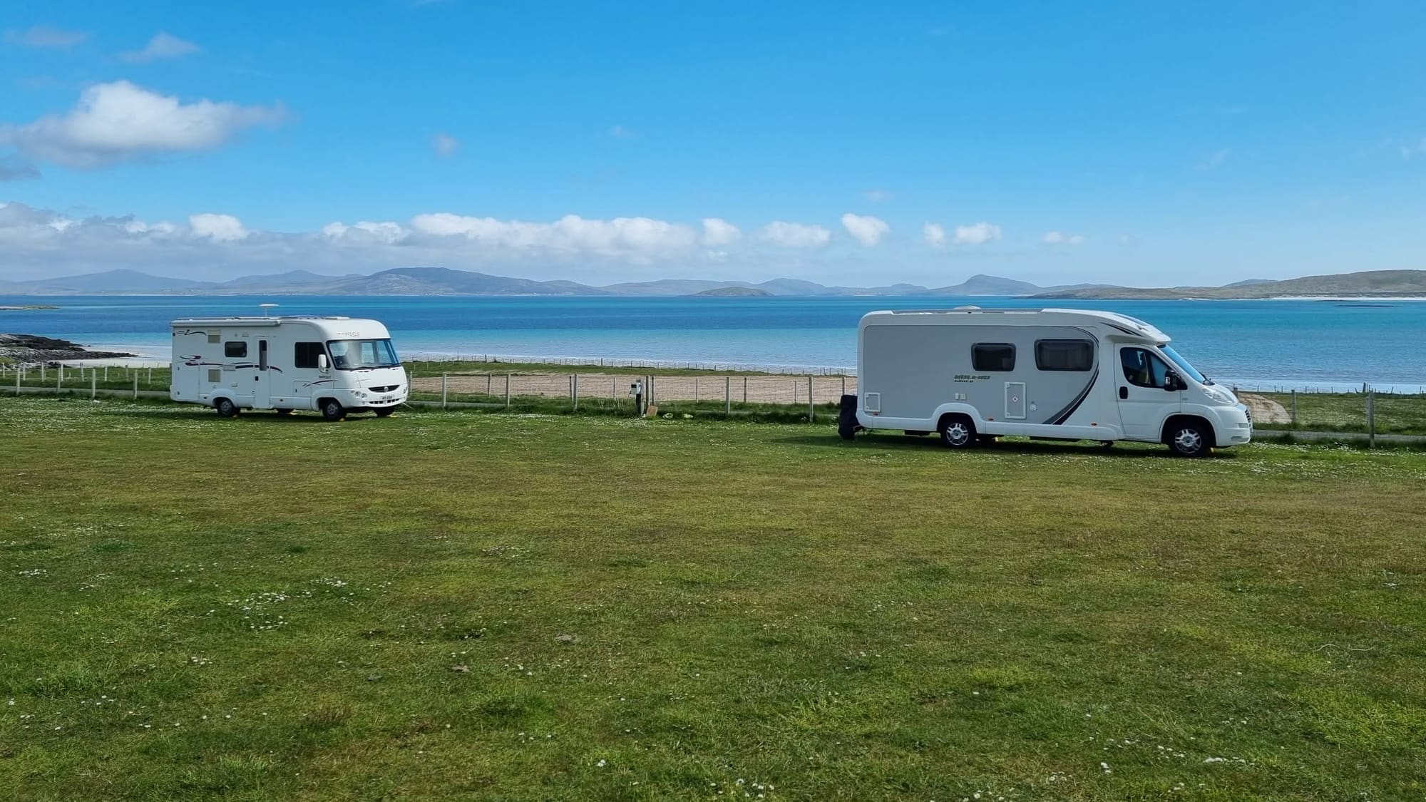 Two motorhomes parked on a mown grass field behind a wire fence, with a white beach, a turquoise bay and distant hills beyond.