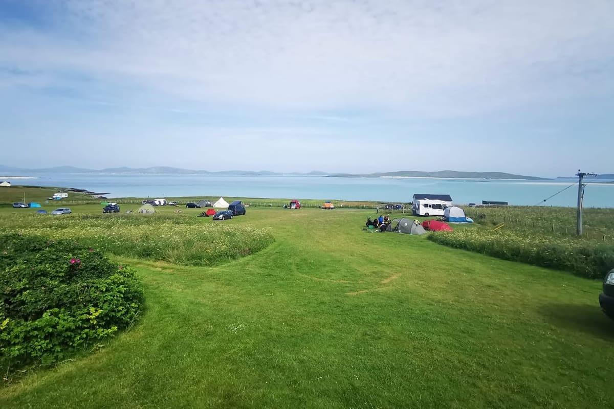 A summer camping field with mown paths through long grass, small tents, campervans and a motorhome, and the sea beyond.