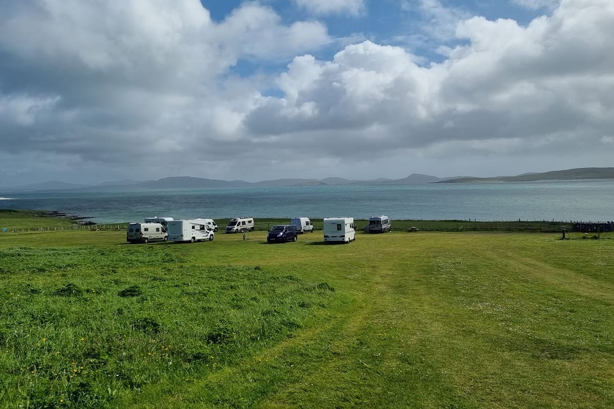 A row of campervans and motorhomes parked along a fence at the edge of a grass field, open sea and hills under a big cloudy sky.