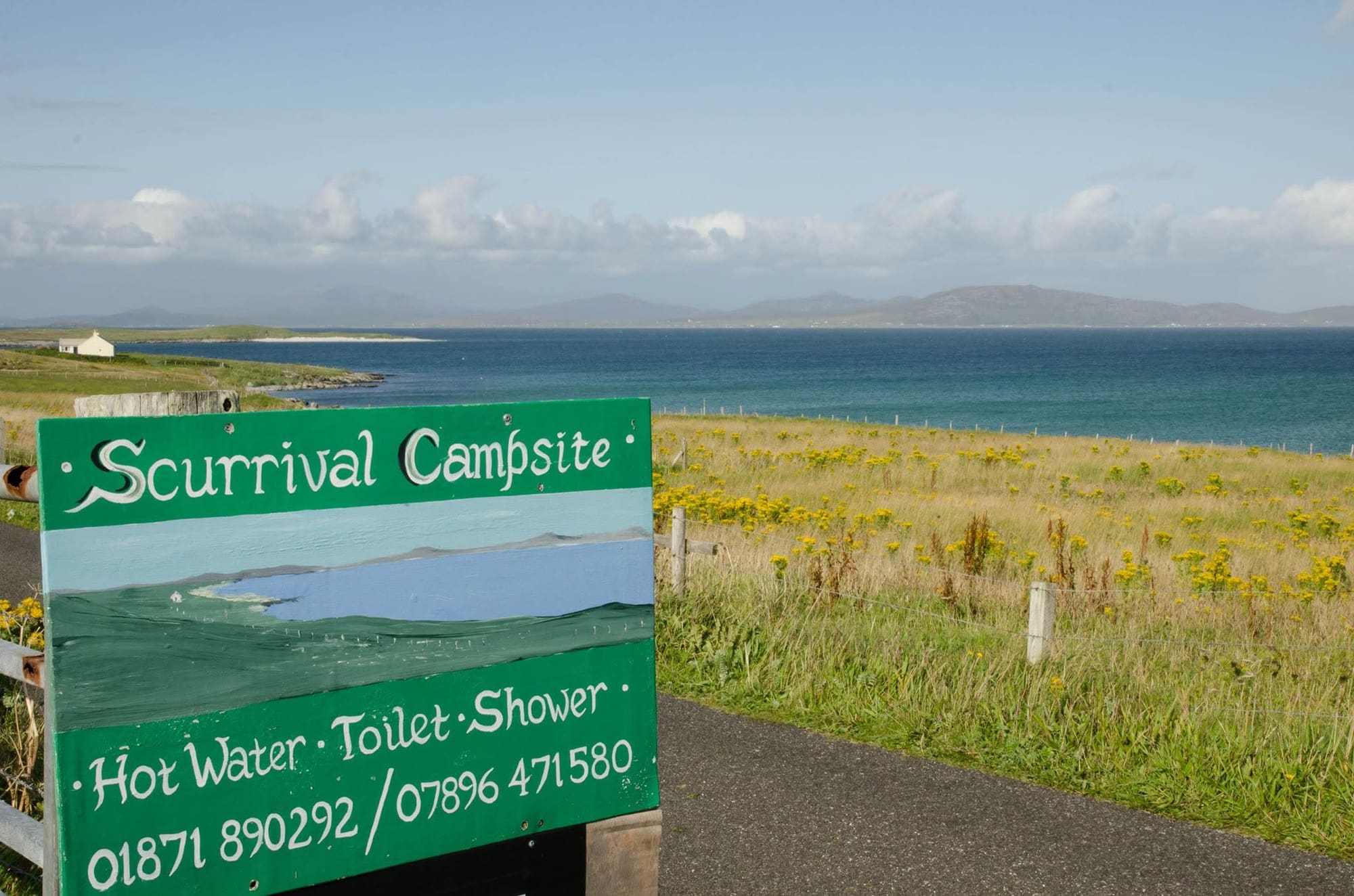 Green hand-painted roadside sign reading "Scurrival Campsite, Hot Water, Toilet, Shower" with the numbers 01871 890292 and 07896 471580, the sea behind it.