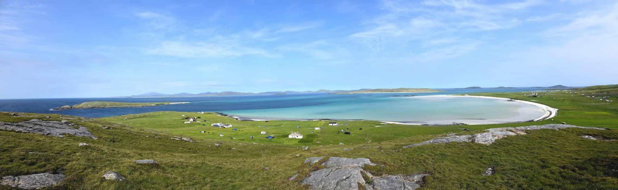 A wide panorama of a crescent white beach and turquoise bay with scattered houses on green land.