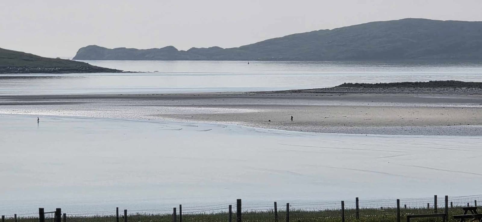 Hazy, silvery low-tide sands with two distant walkers and a dark hill across the water, a wire fence in the foreground.