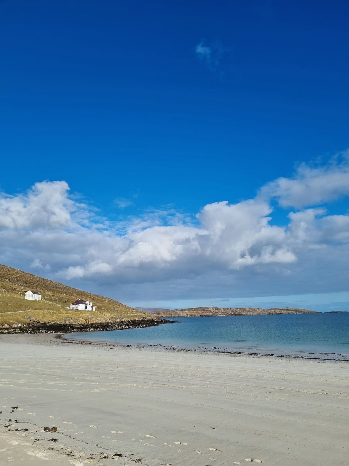A wide white beach and calm blue water under a deep blue sky, with two white cottages on a brown hillside.